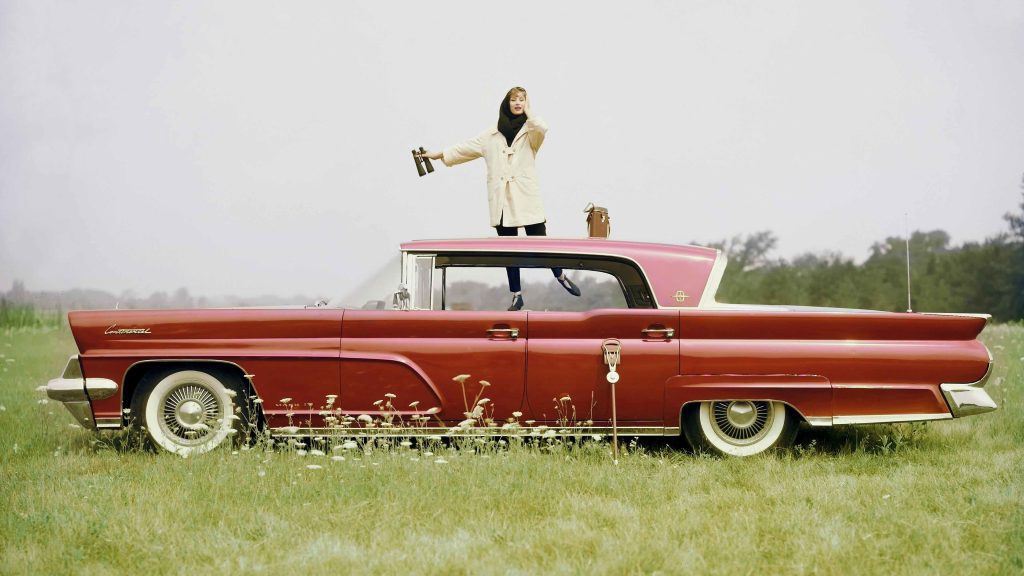Manekenka stoji na rdečem avtomobilu Continental Mark IV hard-top sedan iz leta 1959, parkiranem na travniku z belimi cvetovi. Fotografiral John Rawlings.